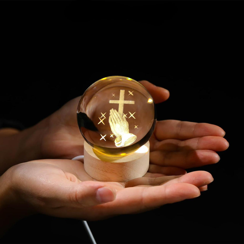 Hand holding a small glass ball with cross and prayer hands design, labeled 'Cross Gifts'.