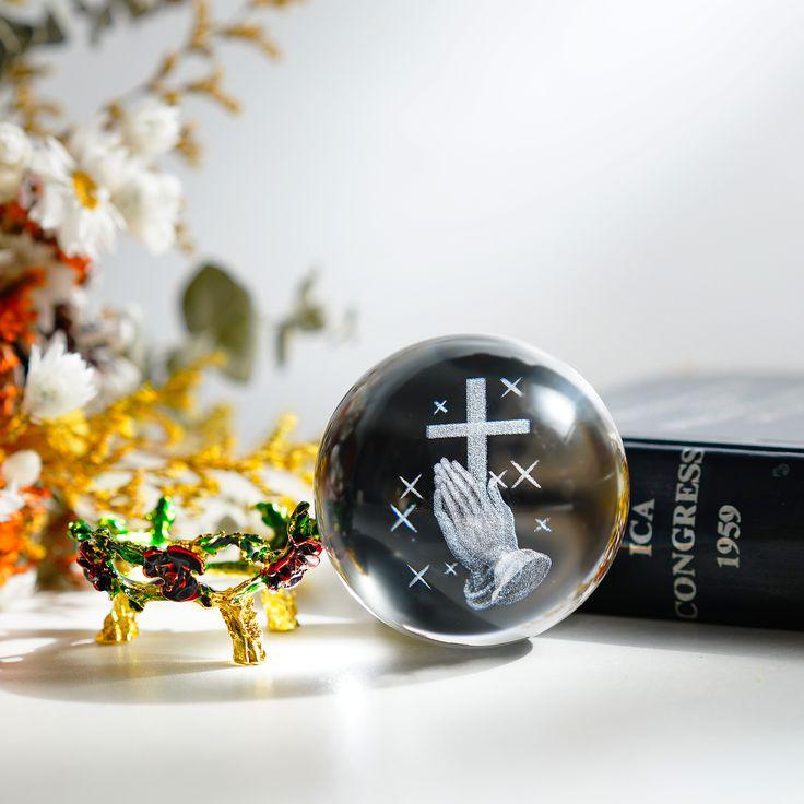Clear glass paperweight with praying hands and cross design on a surface with decorative elements.